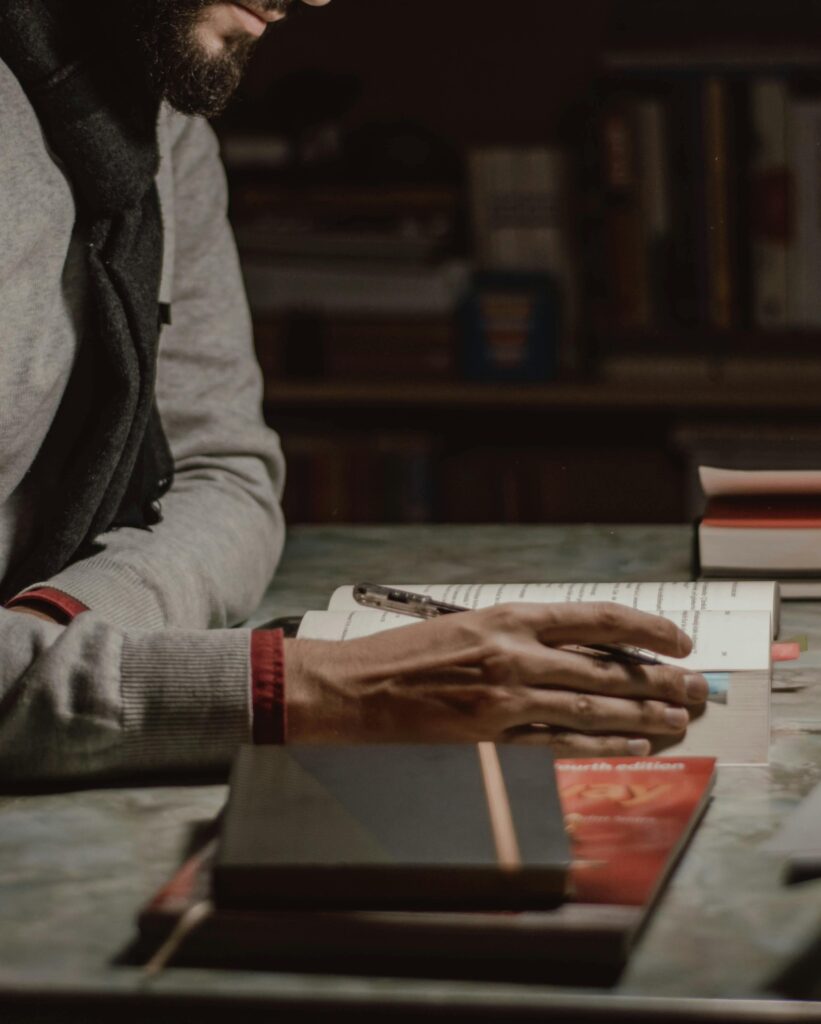 A man deeply absorbed in reading and writing at a dimly lit desk filled with books.