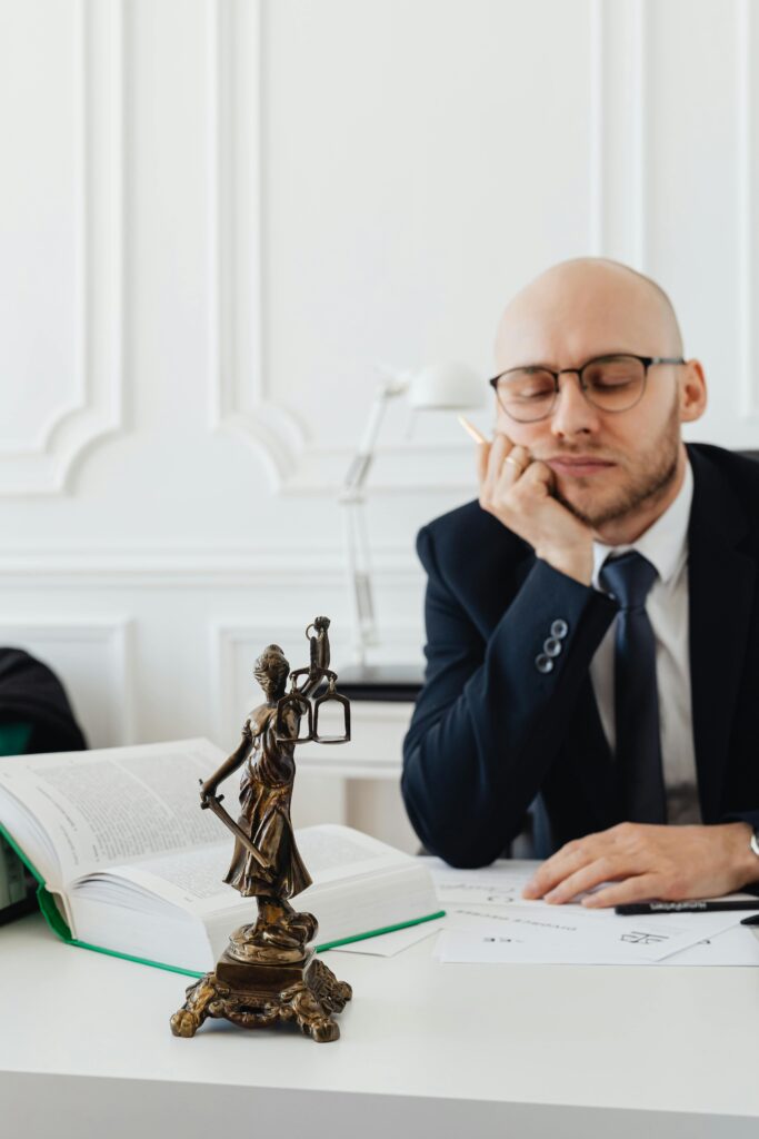A thoughtful lawyer sits at his desk beside a statue of Lady Justice, conveying contemplation.